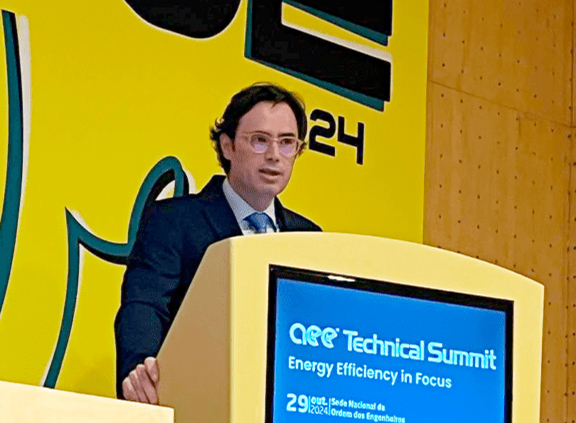 Nelson Lage de fato escuro e óculos fala num púlpito amarelo durante o AQE Technical Summit, com o tema “Energy Efficiency in Focus”. O ecrã embutido no púlpito mostra a data do evento: 29 de Outubro de 2024, realizado na Sede Nacional da Ordem dos Engenheiros. O fundo tem o logótipo grande do evento com fundo amarelo vibrante.