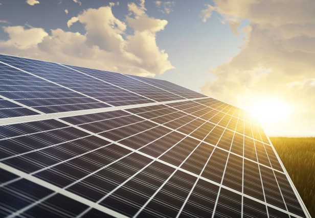 Painel solar fotovoltaico em primeiro plano, captado ao ar livre sob um céu parcialmente nublado. O sol está a nascer ou a pôr-se no horizonte, refletindo luz sobre a superfície do painel, com tons quentes a dominar a cena.