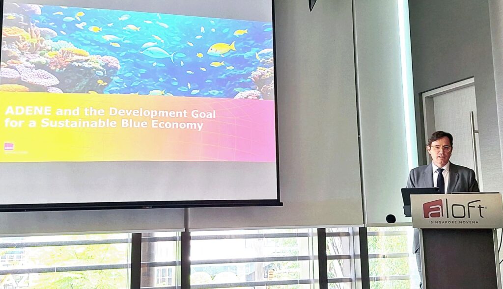 Fotografia de uma apresentação no congresso "Ocean 2024", em Singapura. O orador, em fato e gravata, encontra-se ao púlpito com o logótipo "Aloft Singapore Novena". No ecrã é visível o slide com o título: “ADENE and the Development Goal for a Sustainable Blue Economy”, acompanhado por uma imagem colorida de um recife de coral com peixes tropicais.