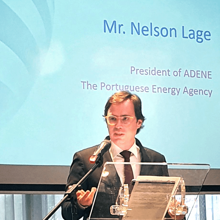 Fotografia de Nelson Lage, Presidente da ADENE – Agência para a Energia, a discursar num púlpito com microfone, durante uma conferência. Atrás dele, um ecrã de projeção exibe o seu nome e título em inglês: “Mr. Nelson Lage, President of ADENE, The Portuguese Energy Agency”. A sala tem iluminação natural vinda de uma janela coberta por cortinas translúcidas.