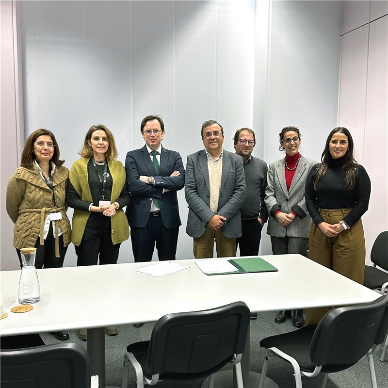 Um grupo de sete pessoas está posicionado lado a lado em frente a uma parede branca. Estão todos vestidos com roupas formais ou de trabalho. À frente do grupo há uma mesa branca com duas garrafas de vidro e alguns documentos pousados. Todos olham para a câmara com expressões neutras ou sorridentes.